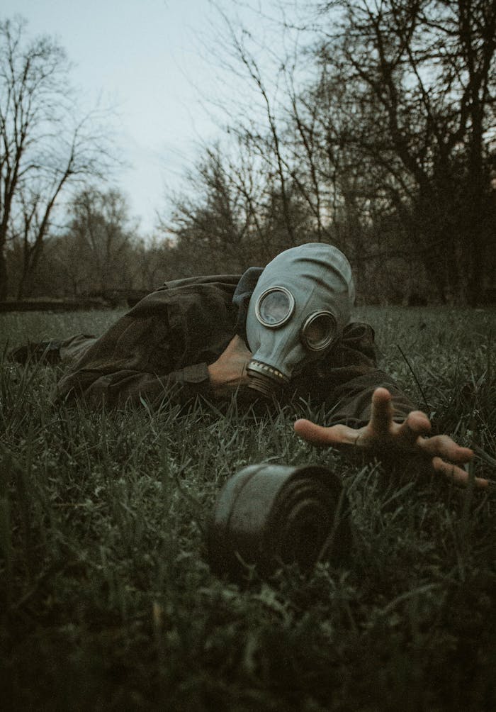 A person wearing a gas mask reaches across the grass in a moody forest setting.